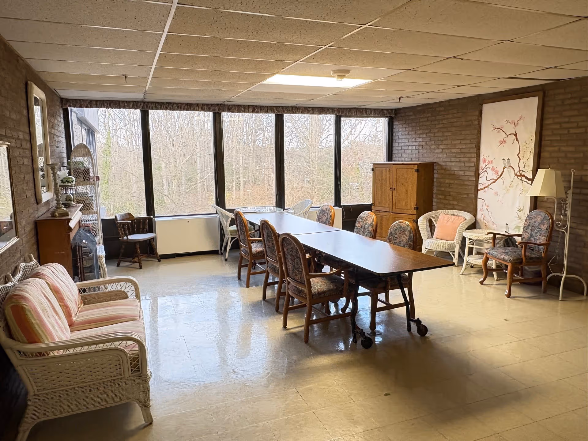 A spacious room with large windows showing a view of leafless trees outside. Inside, there is a long table with several wooden chairs around it, a white wicker loveseat with striped cushions, a wooden cabinet, a floor lamp, and various other chairs and small tables. The walls are brick, and the floor is a light-colored tile.