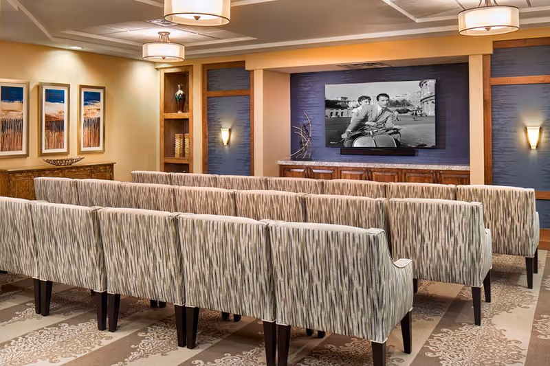 A cozy media room with multiple rows of patterned upholstered chairs facing a wall-mounted flat-screen TV displaying a black and white movie scene. The room features warm lighting, decorative wall sconces, framed artwork, and built-in wooden shelves with decorative items.