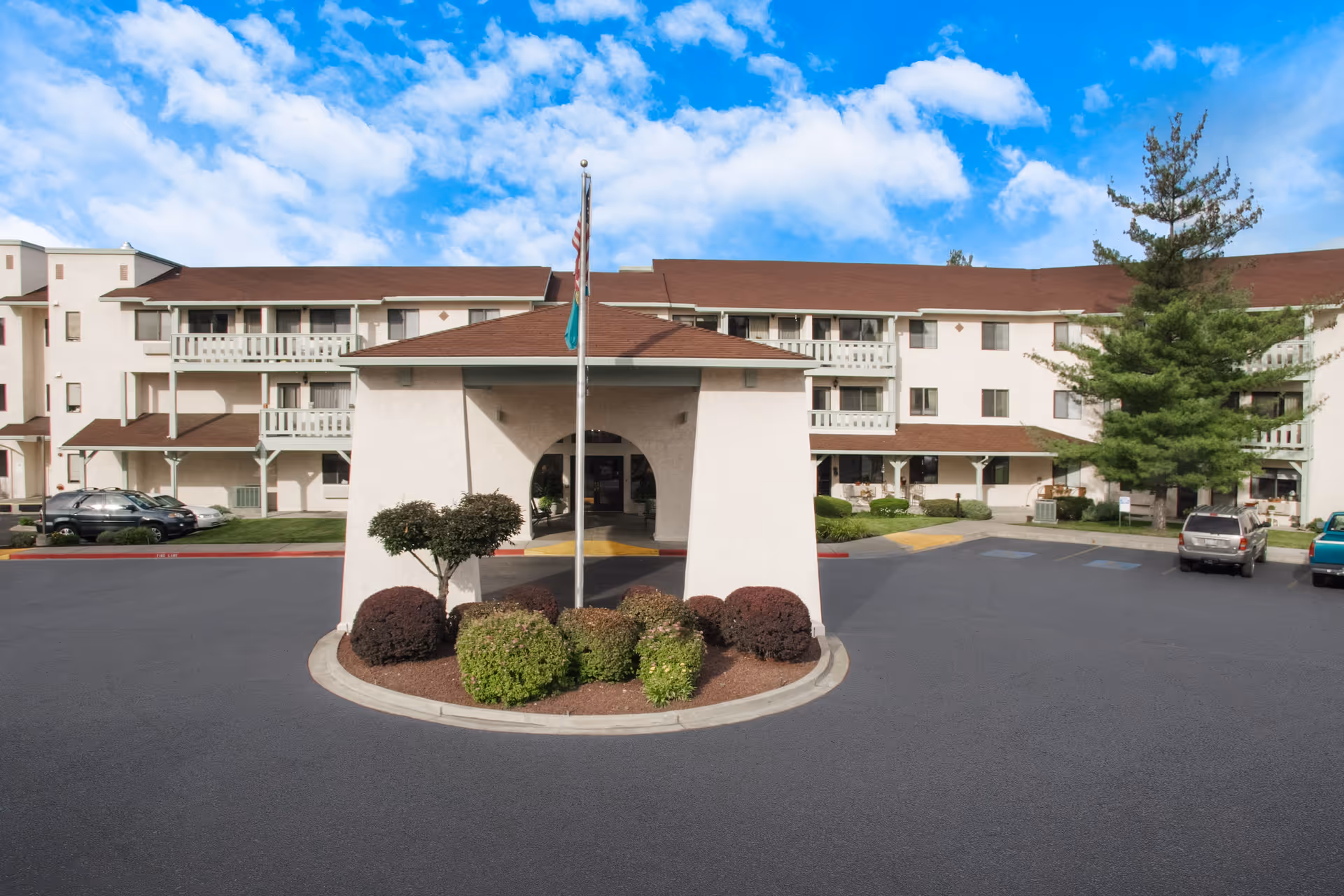 Front exterior view of a multi-story senior living facility with a covered entrance, landscaped bushes, and an American flag on a flagpole. Several cars are parked in the parking lot, and the sky is partly cloudy.