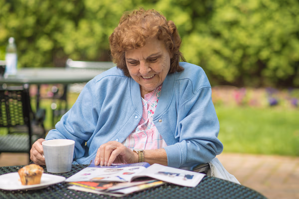 An elderly woman with curly hair wearing a light blue cardigan sits at an outdoor metal table, smiling while looking at a magazine. On the table, there is a white cup and a plate with a muffin. The background shows green bushes and a garden area.