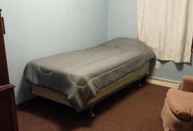 Single bed with a gray comforter in a small bedroom with light blue walls, a curtain, and an upholstered chair.