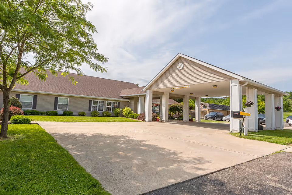 Exterior view of Riverside Landing Skilled Nursing & Rehabilitation showing a covered entrance with white pillars, a driveway, green lawn, trees, and a building with beige siding and brown roof under a partly cloudy sky.