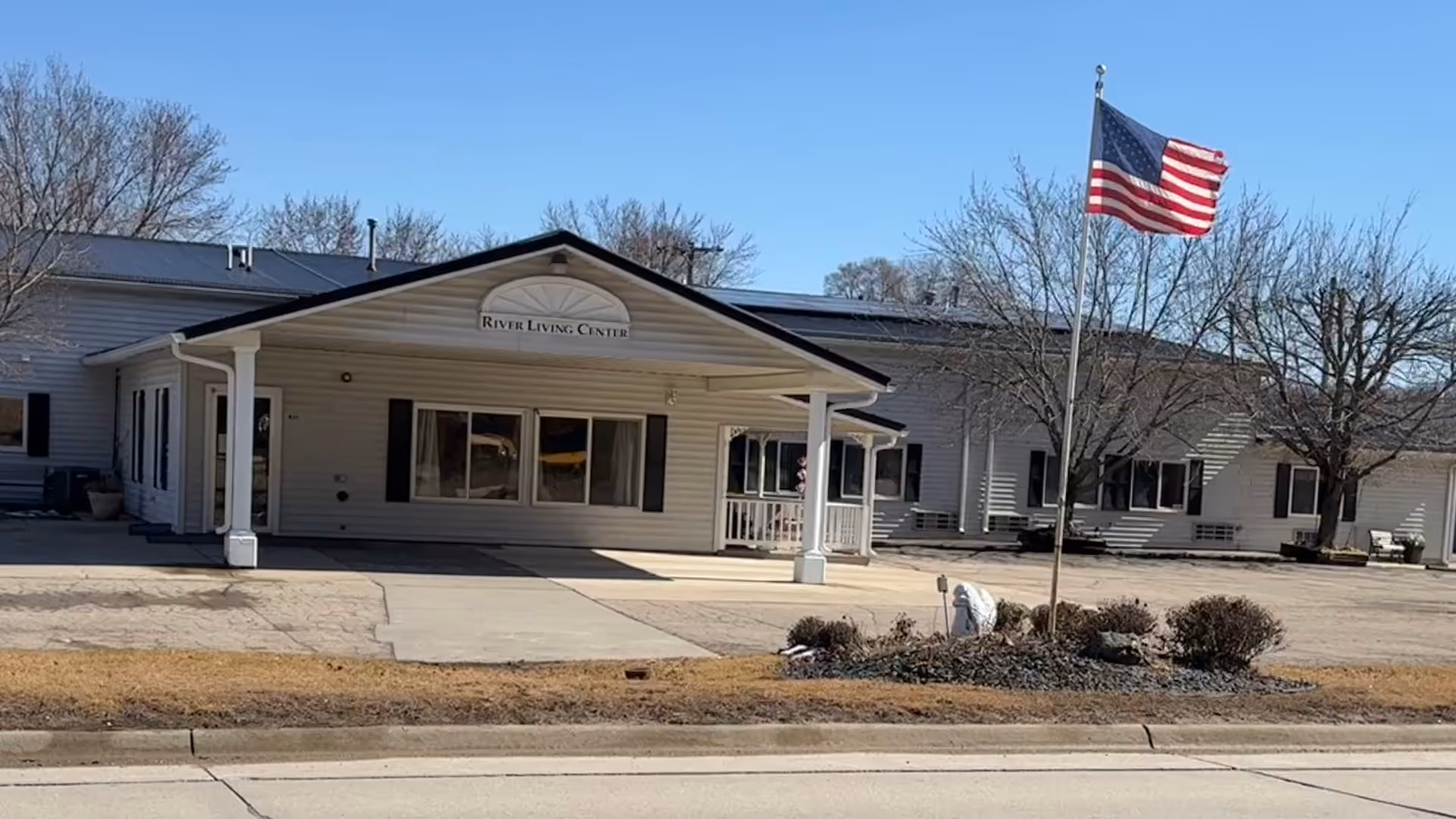 Front entrance of a single-story senior living facility with a covered portico, landscaping, and an American flag, showing a sign that reads "River Living Center."