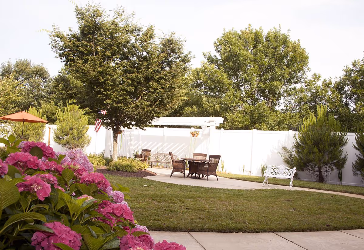 Sunlit outdoor courtyard with pink hydrangeas in the foreground, a patio table and chairs, white fence, and trees.