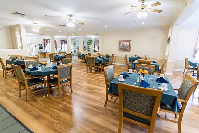 A spacious dining room with multiple wooden tables covered with teal tablecloths, each set with white cups, glasses, silverware, and napkins. The room has wooden flooring, ceiling fans, and light-colored walls with framed artwork. Large windows and glass doors allow natural light to enter the space.