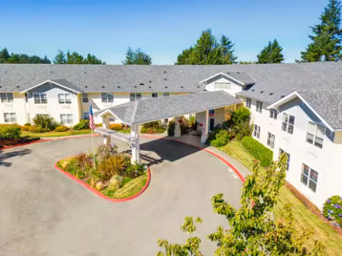 Front exterior of a two-story U-shaped senior living building with a covered porte-cochère, driveway, and landscaped grounds.