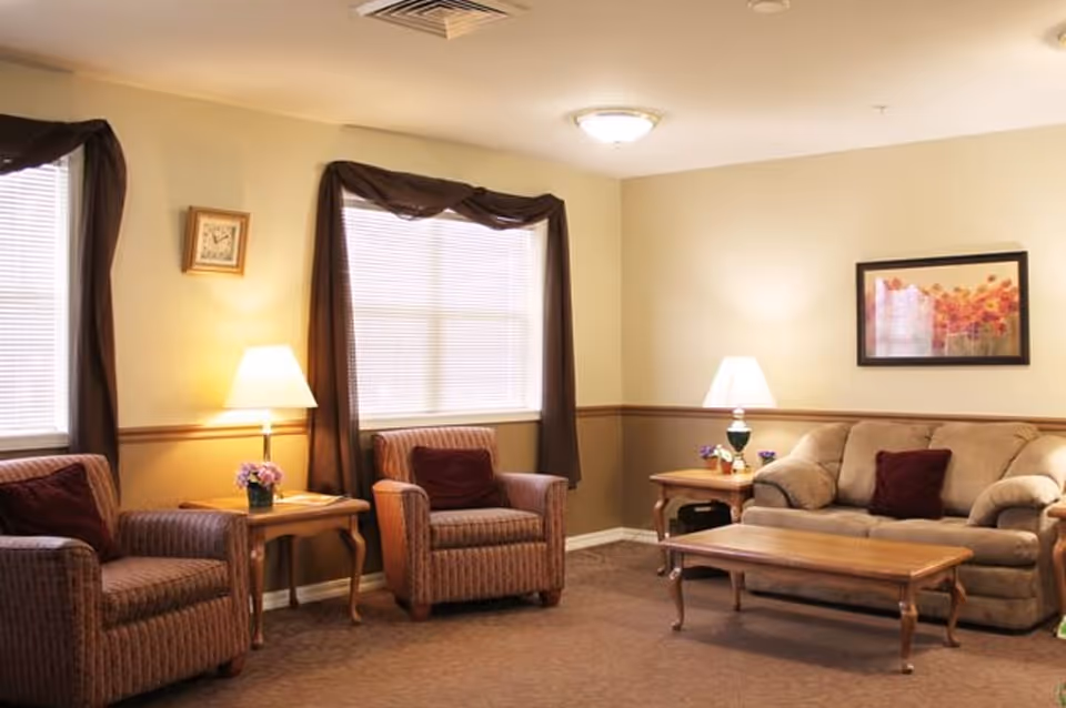 A cozy living room with two striped armchairs and a beige sofa, each with a dark red cushion. There are two wooden side tables with lamps and small flower arrangements. The room has beige walls with a wooden chair rail, two windows with brown curtains, a framed floral picture on the wall, and a wooden coffee table in the center.
