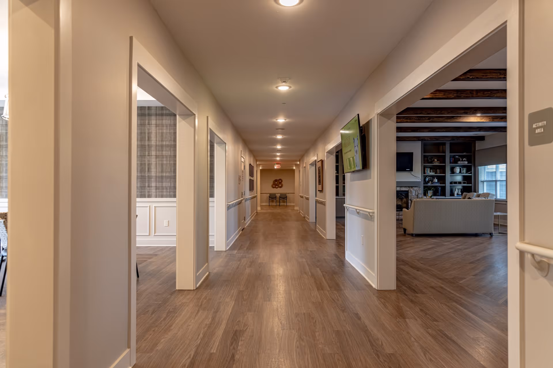Well-lit interior hallway with open doorways revealing a lounge and activity areas in a senior living facility.