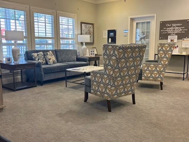 A senior living facility common area with a gray sofa adorned with patterned cushions, two patterned armchairs, a white marble coffee table, side tables with lamps, and large windows with white blinds letting in natural light. A mission statement is displayed on the wall in the background.