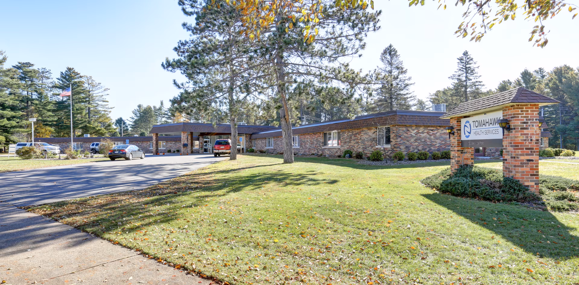 Exterior view of Tomahawk Health Services building with a brick facade, surrounded by green grass and trees. Several cars are parked near the entrance, and an American flag is visible on a flagpole to the left.