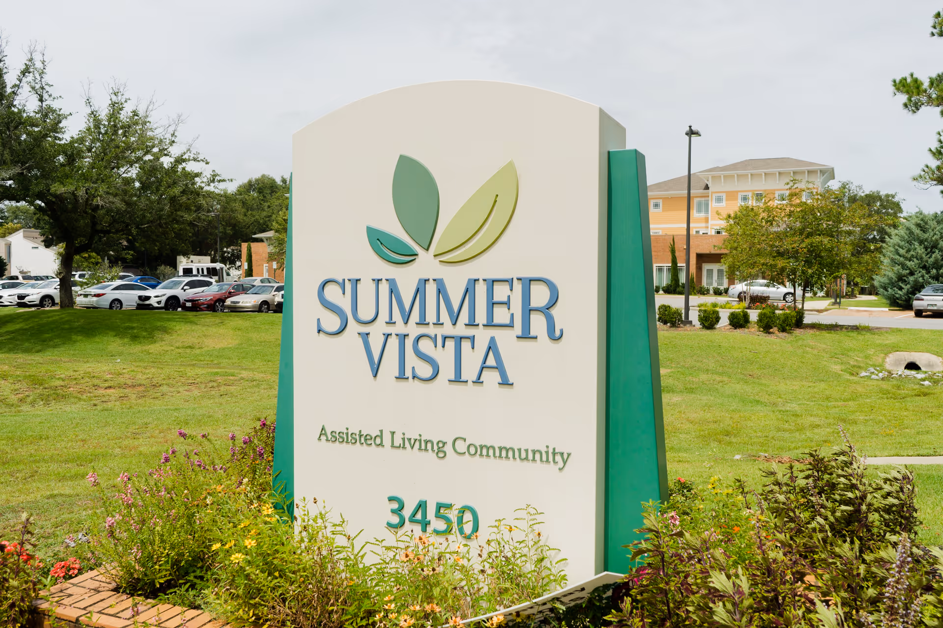 Entrance sign reading 'SUMMER VISTA Assisted Living Community 3450' on a landscaped lawn with the facility building and parked cars in the background.