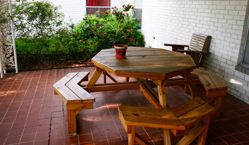 A wooden octagonal picnic table with attached benches and a wooden chair on a tiled patio. There is a small decorative pot on the table. The patio is adjacent to a white brick wall with a window and surrounded by green bushes and plants.