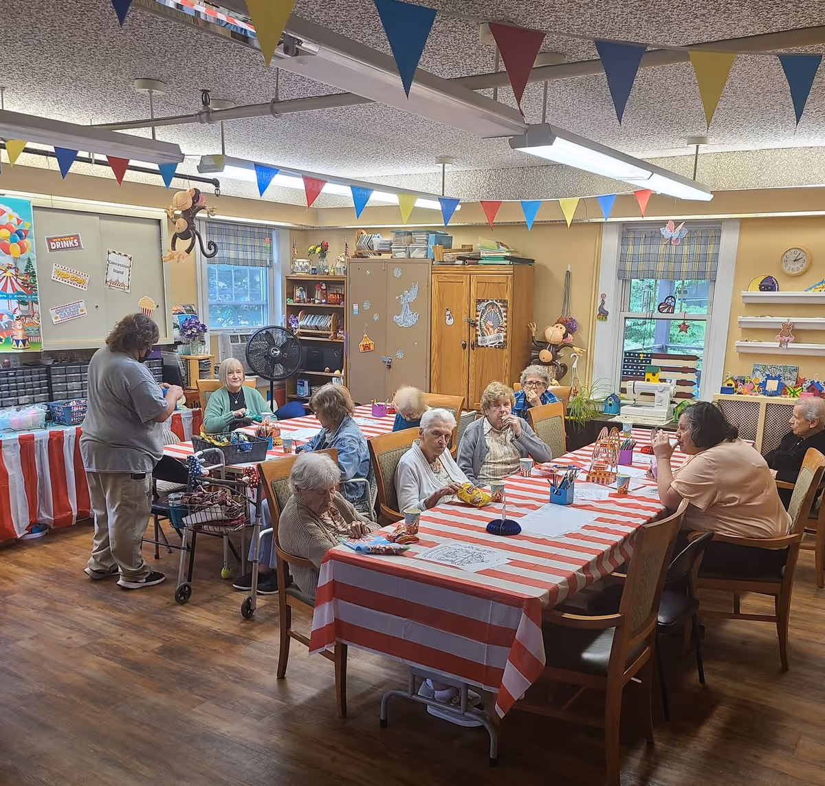 A group of elderly residents seated around a long table with red-and-white striped tablecloths in a decorated activity/dining room.