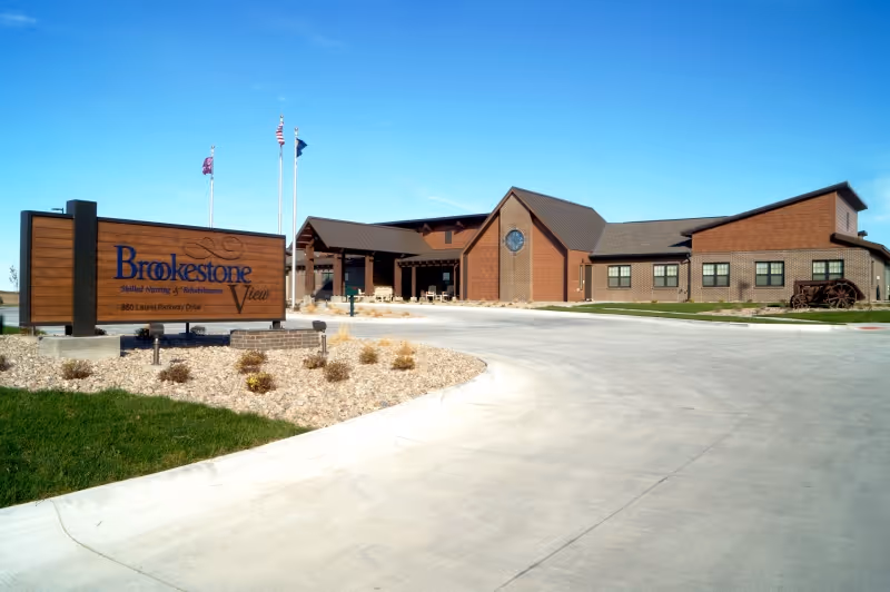 Exterior view of Brookestone View, a senior living and rehabilitation facility, showing a large brick building with a covered entrance, a curved driveway, a landscaped area with small bushes, and a wooden sign with the facility's name and address. Three flagpoles with flags are visible behind the sign under a clear blue sky.