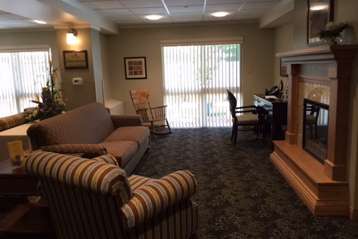 A cozy living room with a patterned carpet, a striped armchair, a brown sofa, a wooden rocking chair, a desk with a chair, and a wooden fireplace. Large windows with vertical blinds allow natural light into the room.