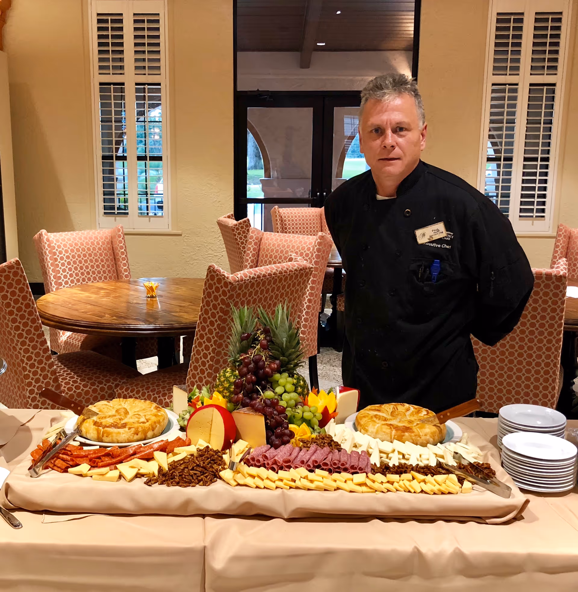 A chef in a black uniform stands behind a buffet table arranged with cheeses, fruits, nuts and pastries in a dining room.