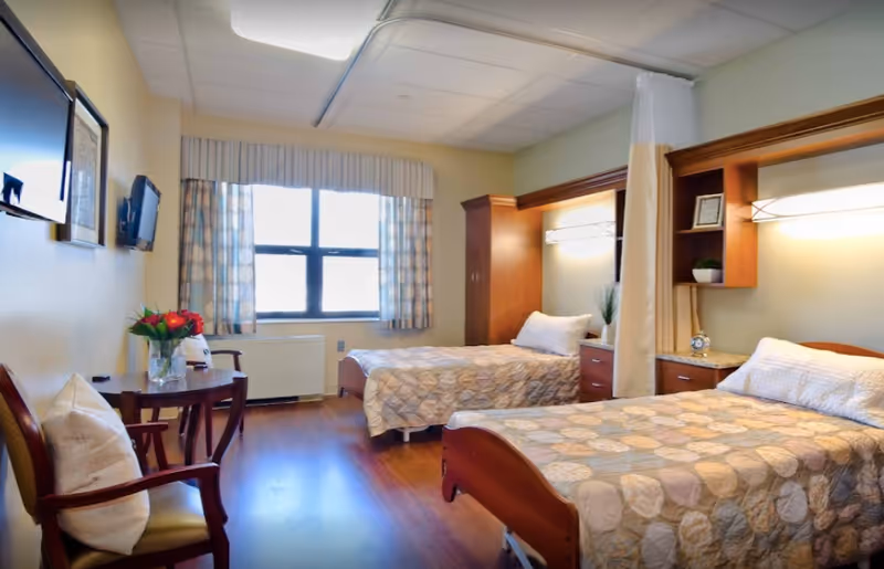 A bright and clean bedroom in a senior living facility with two single beds, each with patterned bedspreads and white pillows. The room has wooden furniture including a small table with a vase of red flowers and two chairs with cushions. There is a window with patterned curtains letting in natural light, a wall-mounted TV, and built-in wooden cabinets with lighting above the beds. A privacy curtain is partially drawn between the beds.