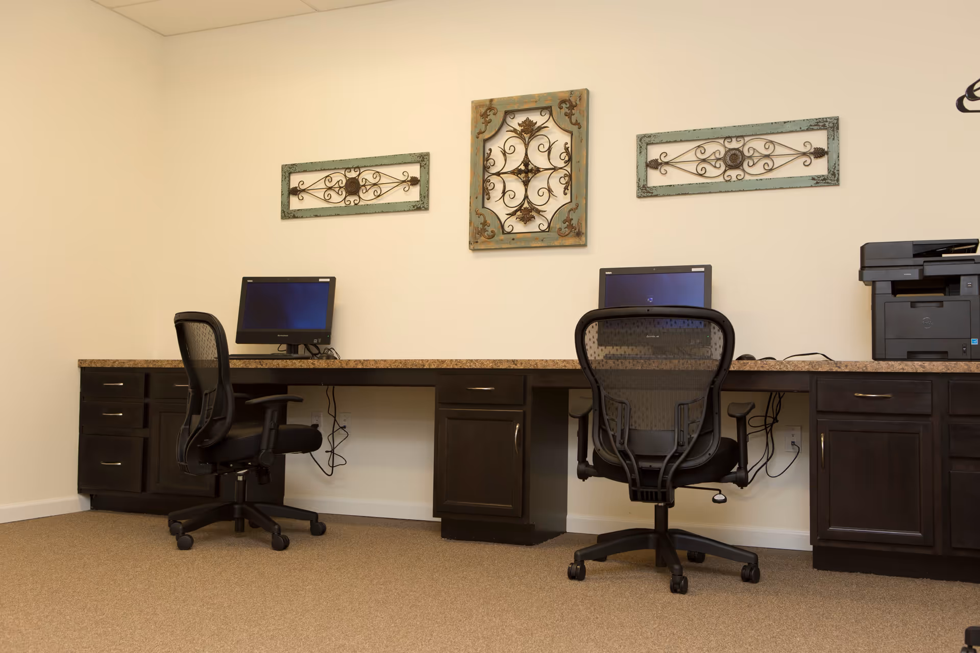 A small office area with two black mesh office chairs in front of a long dark wood desk with a granite countertop. Two computer monitors are placed on the desk, and a printer is on the right side. Three decorative metal wall hangings are mounted on the beige wall above the desk.
