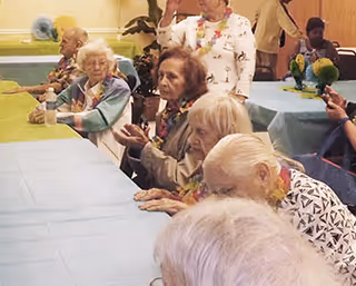 A group of elderly people sitting around tables covered with blue tablecloths in a communal indoor setting, some wearing leis and engaging in conversation or activities.