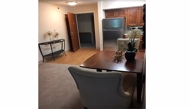 A dining area with a wooden table and upholstered chairs opens to a small kitchen with a stainless-steel refrigerator and an open doorway.