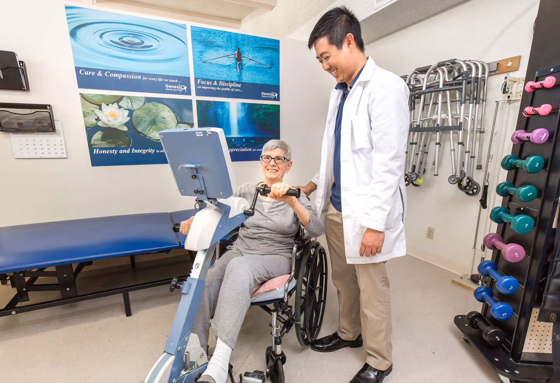 An elderly woman in a wheelchair is using a hand cycle exercise machine with the assistance of a smiling male healthcare professional in a white coat. The room has exercise equipment including dumbbells and walkers, and motivational posters on the wall.