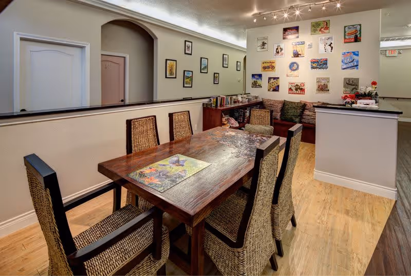 A cozy dining area with a wooden table surrounded by six wicker chairs. The table has a colorful placemat and a puzzle in progress. The room features light-colored walls adorned with various framed pictures and vintage-style posters. There is a cushioned bench with pillows along the far wall and a small bookshelf with books and decorative items. The flooring is a combination of light and dark wood tones.