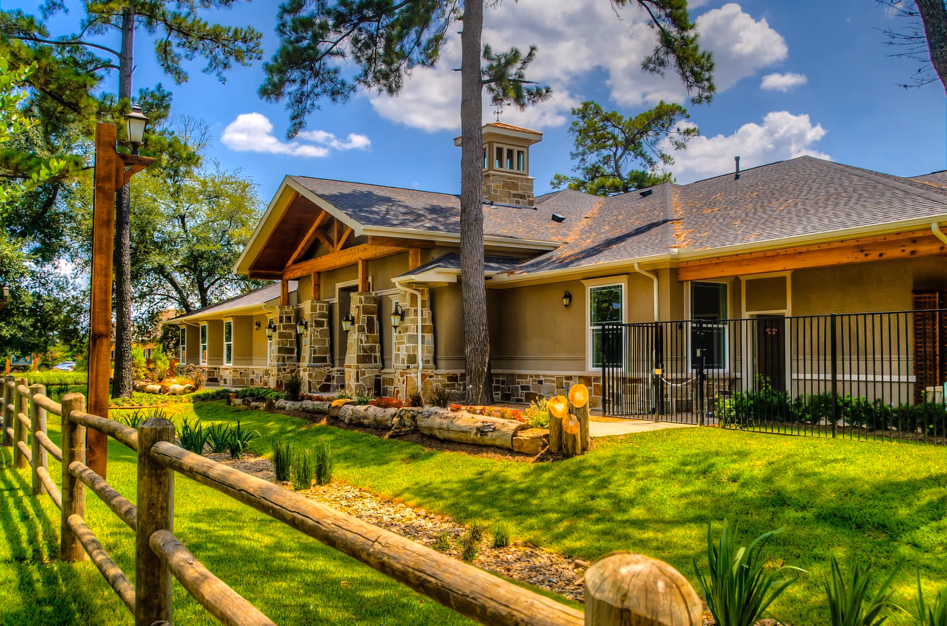 Exterior view of a single-story building with stone and beige siding, surrounded by green grass, trees, and a wooden fence under a partly cloudy blue sky.