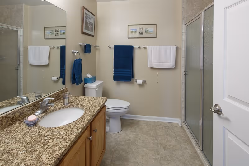 A clean bathroom with a granite countertop sink, wooden cabinets, a toilet, and a glass-enclosed shower. The walls are beige with two towel racks holding blue and white towels. There are two framed pictures on the wall above the toilet.