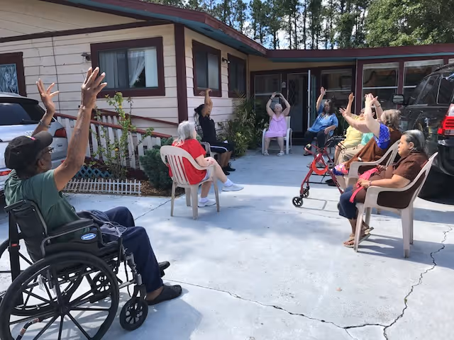 A group of elderly individuals seated in a semi-circle outdoors on a concrete patio in front of a single-story building. Some participants are in wheelchairs or using walkers, and they are raising their arms, possibly engaging in a group exercise or activity. The setting is sunny with trees visible in the background.