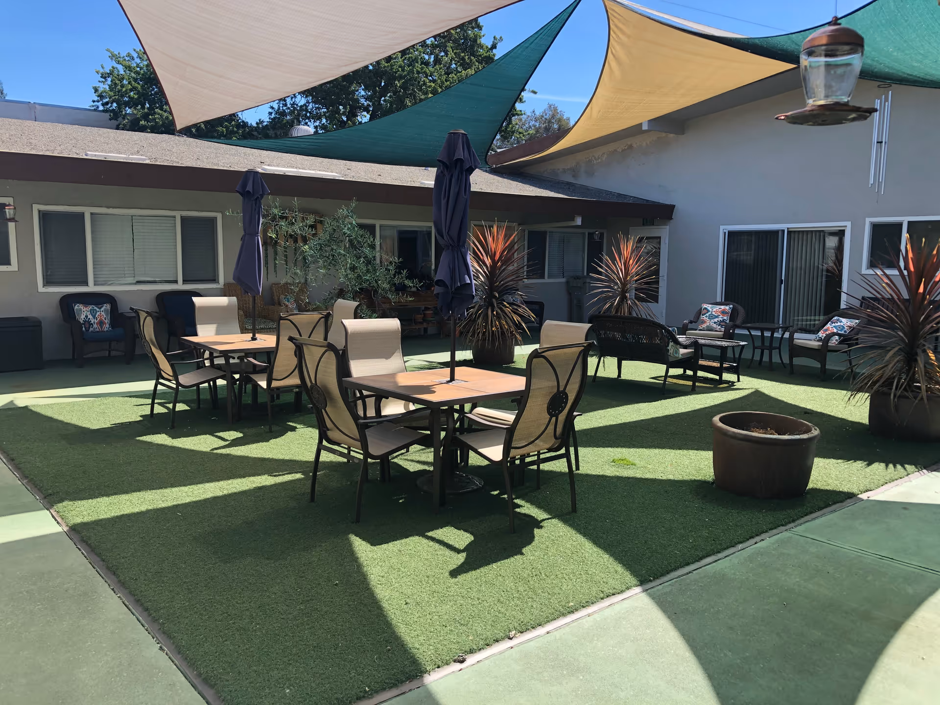 Outdoor courtyard area with artificial grass, several tables with chairs and closed umbrellas, lounge chairs with cushions, potted plants, and shade sails overhead providing partial shade.