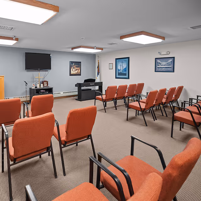 A small meeting or chapel room with rows of red upholstered chairs facing a TV, podium, and electric keyboard.