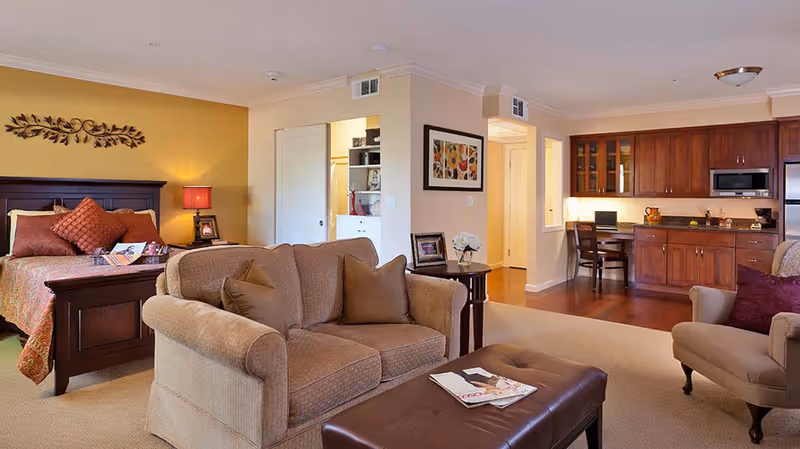 A spacious senior living suite featuring a cozy living area with a beige sofa, armchair, and a brown leather ottoman. The background shows a bedroom with a wooden bed frame, orange and brown bedding, and a bedside table with a lamp. To the right, there is a kitchenette with wooden cabinets, a countertop, and a small desk with a chair. The room has warm lighting and a welcoming atmosphere.