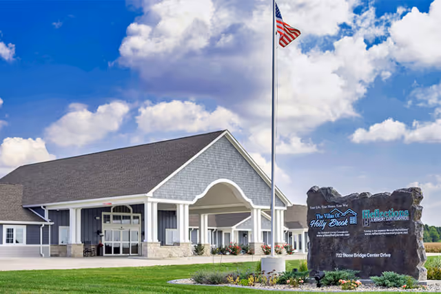 Exterior view of Villas of Holly Brook Assisted Living & Memory Care building in Rantoul, IL, featuring a large covered entrance with white columns, a manicured lawn, an American flag on a tall flagpole, and a large stone sign displaying the facility's name and address under a partly cloudy blue sky.