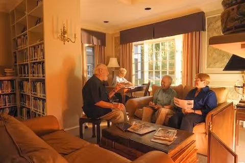 Four older adults sit and converse in a warmly lit living room with bookshelves, a coffee table, and large windows.
