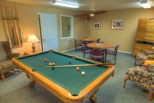 A recreational room with a pool table in the center, several billiard balls and a cue stick on the table. The room also contains a few round tables with purple chairs, a wooden cabinet, floral upholstered armchairs, a beige armchair, a lamp on a side table, and framed pictures on the walls.