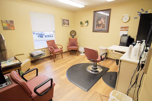 A small room with light yellow walls and wood flooring, furnished with several chairs including a salon chair on a black mat, a small table with hair care products, a TV, and wall decorations including framed pictures and a clock. A window with a white blind lets in natural light.