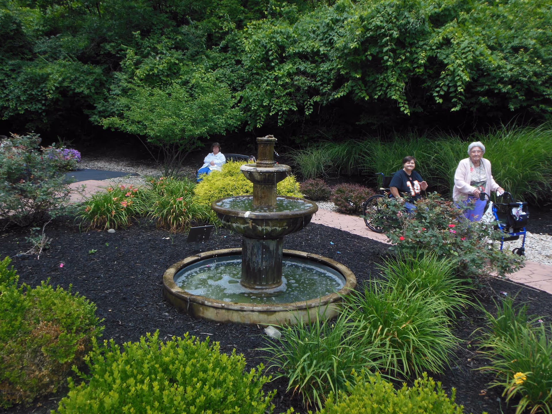 A three-tiered stone fountain in a landscaped garden with shrubs and two people on a path, one in a wheelchair and one using a walker.