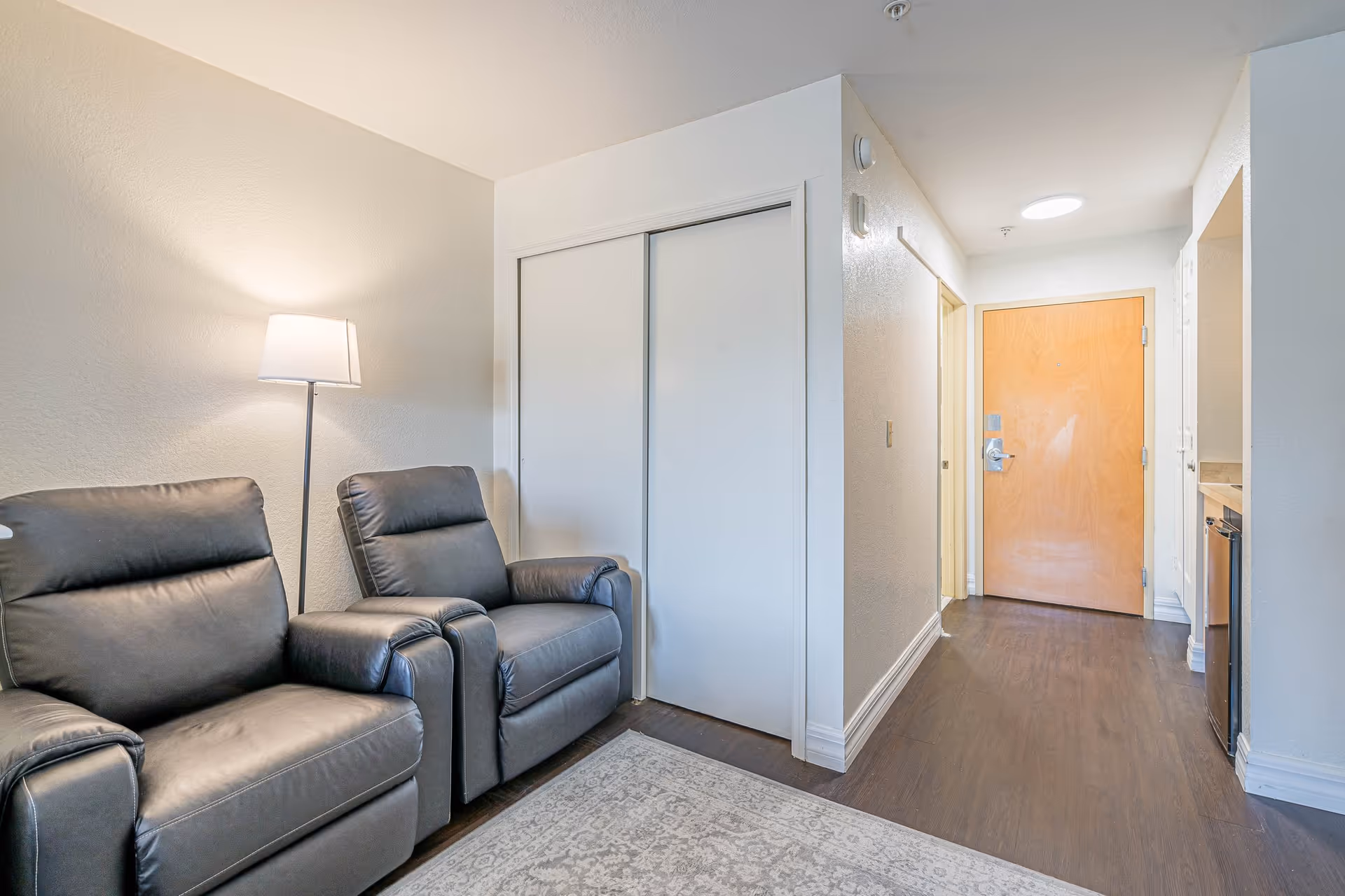 Small living area with two gray leather recliners beside a floor lamp, a closet with sliding doors, and a hallway leading to a wooden entrance door.