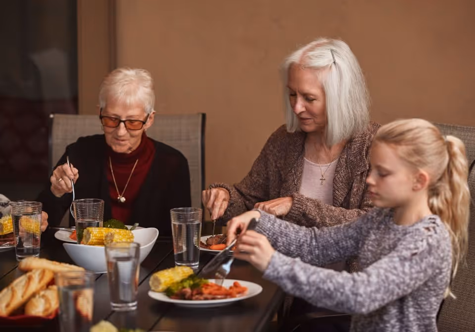 Two elderly women and a young girl sitting at a dining table, eating a meal that includes corn on the cob, carrots, and bread, with glasses of water on the table.