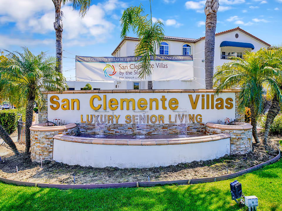 Outdoor view of the entrance sign for San Clemente Villas Luxury Senior Living, featuring a stone and tile structure with water fountains, surrounded by palm trees and greenery, with a building and a banner in the background.