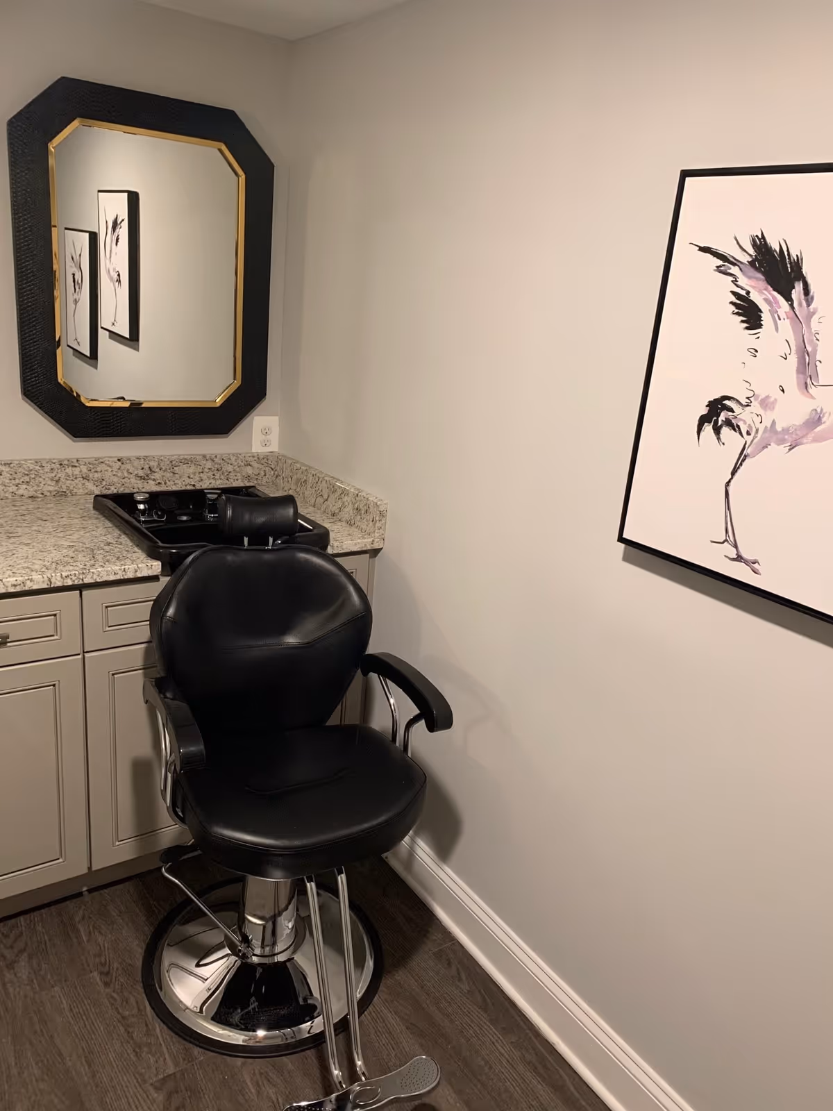 A small hair salon area with a black salon chair in front of a granite countertop with a built-in black sink. Above the countertop is a black-framed mirror reflecting two pieces of wall art. On the adjacent wall, there is a framed painting of a bird with black and purple accents. The floor is dark wood, and the walls are painted light gray.