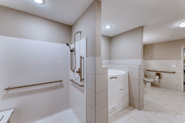 A spacious, accessible bathroom featuring a walk-in shower with grab bars and a handheld showerhead, a walk-in bathtub with a door, and a toilet with grab bars. The room has beige walls, white tiled floors, and bright ceiling lights.