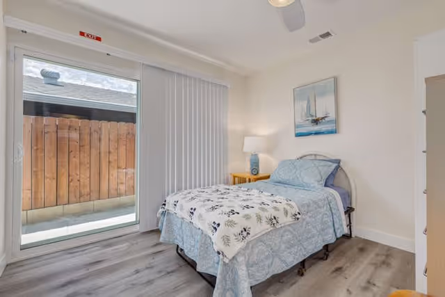 A bright bedroom with a single bed covered in blue and white bedding, a wooden nightstand with a lamp, a painting of a sailboat on the wall, a ceiling fan, and a sliding glass door with vertical blinds leading to an outdoor area with a wooden fence.