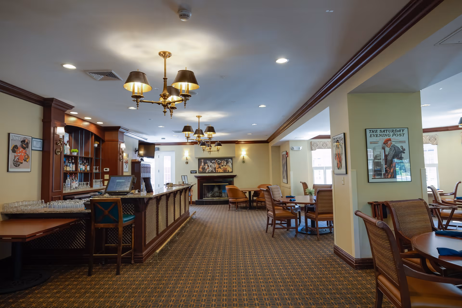 A spacious and well-lit common area with a bar counter on the left side, wooden chairs, and tables arranged for seating. The room features patterned carpet flooring, multiple chandeliers with lampshades, framed vintage posters on the walls, and a fireplace at the far end. Large windows allow natural light to brighten the space.