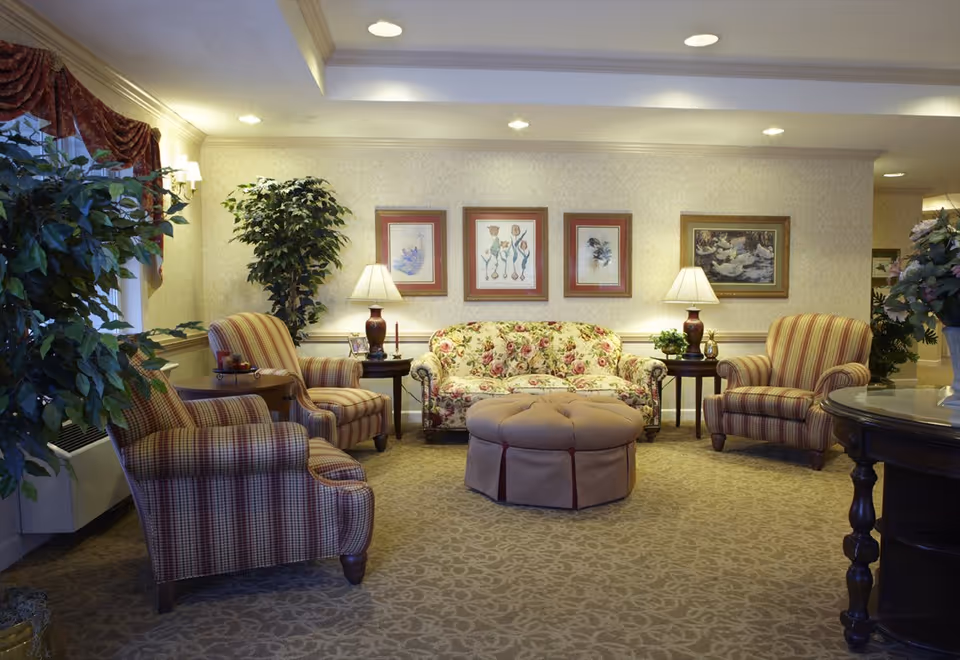 A cozy living room area with a floral patterned sofa, two striped armchairs, a round tufted ottoman in the center, two side tables with lamps, framed artwork on the wall, and several large potted plants. The room has warm lighting and carpeted flooring.