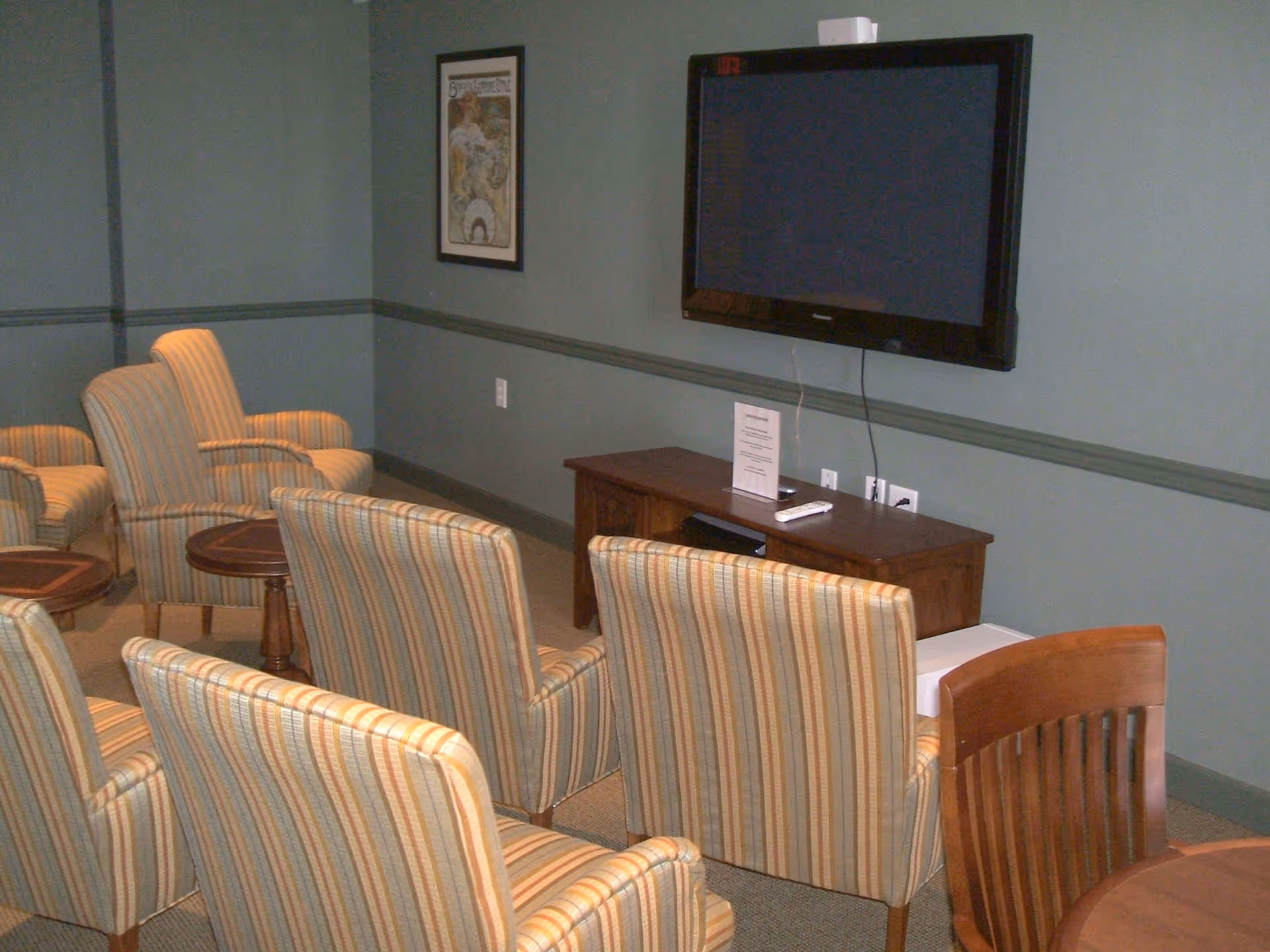 A small lounge or common area with several striped upholstered armchairs arranged facing a wall-mounted flat screen TV. Below the TV is a wooden console table with a remote control and a small sign on it. The walls are painted green with a chair rail molding, and there is a framed picture hanging on the wall.