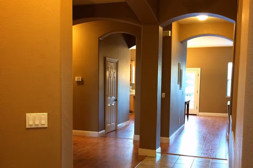 Interior view of a hallway in a senior living facility with warm beige walls, wooden flooring, arched doorways, and a door at the end leading to another room with natural light coming through windows.