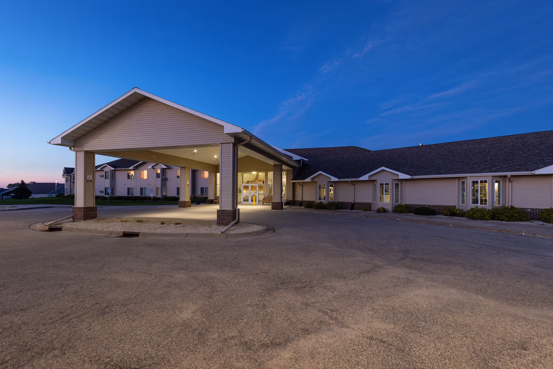 Exterior view of Prairie Meadows Senior Living facility at dusk, showing a large covered entrance with lights on and a multi-story building in the background under a clear blue sky.