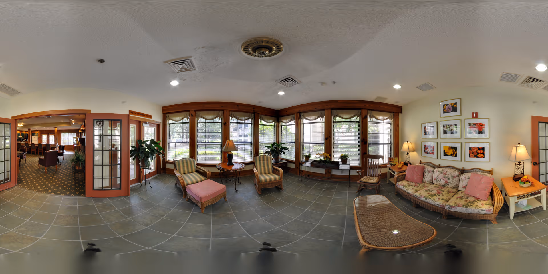A cozy sitting area in a senior living facility with large windows letting in natural light. The room features a floral-patterned sofa with pink cushions, two striped armchairs with a matching ottoman, a wicker coffee table, side tables with lamps, and several potted plants. The walls are decorated with framed floral artwork. Adjacent to this area is a carpeted room with additional seating visible through glass-paneled double doors.