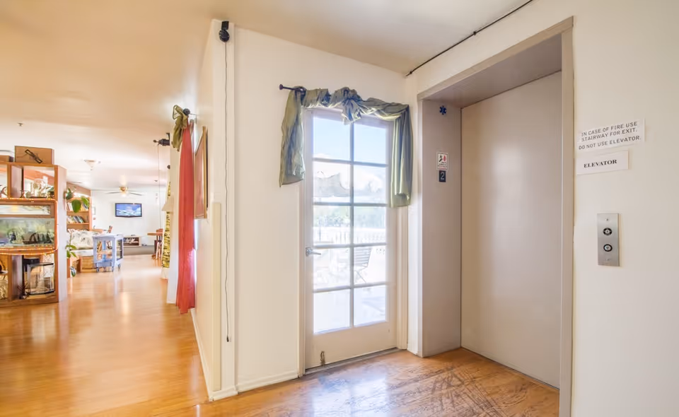 Interior view of a senior living facility hallway with wooden flooring, an elevator door on the right, and a glass door with a green curtain leading to an outdoor patio. The hallway extends to a living area with furniture and a television mounted on the wall.